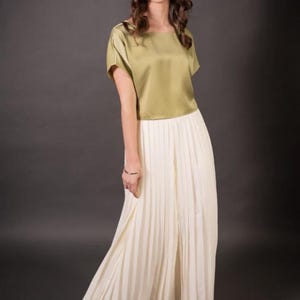 Może przedstawiać: Kobieta w dwuczęściowym stroju. Góra to bluzka z krótkim rękawem z oliwkowozielonego satyny. Dół to długa, plisowana, kremowa spódnica. Strój jest zestawiony ze srebrnymi sandałami. Tło jest ciemnoszare.