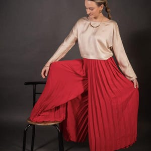 May include: A woman's outfit featuring a long-sleeved, light beige top and wide-legged, pleated red pants. The outfit is paired with gold-colored jewelry and sandals. The woman is sitting on a black stool against a dark gray background.