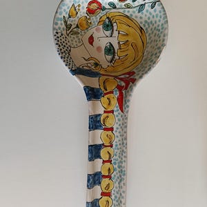 May include: A ceramic spoon rest with a whimsical design. The spoon rest features a blonde-haired girl with large green eyes, red lips, and a red ribbon. The handle has blue and white stripes and a decorative yellow and red design.