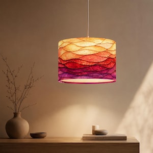 May include: A cylindrical pendant lamp shade featuring a gradient pattern of wavy, overlapping horizontal lines in shades of yellow, orange, red, and purple. The shade is suspended from a single white cord and emits a warm, diffused light.