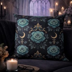 Peut inclure: Coussin carré avec un fond sombre orné de roses turquoise, de croissants de lune dorés et d'ornements dorés. Le coussin est posé sur un canapé gris foncé, avec des bougies et une fenêtre en arrière-plan.