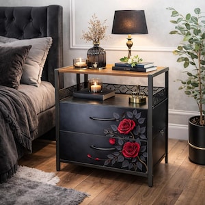 May include: Black nightstand with two drawers, adorned with red rose decals. The nightstand has a metal frame and a wooden top. A black lamp with a gold base sits on top, along with books and candles. A plant in a black pot is to the right.