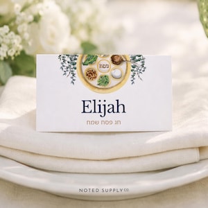 Puede incluir: Una tarjeta de sitio blanca con el nombre "Elijah" en escritura azul marino y texto hebreo debajo. La tarjeta presenta una ilustración de acuarela de un plato de Seder y vegetación. La tarjeta está colocada sobre una servilleta de lino blanca y un plato blanco.