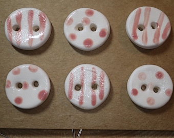 May include: Six white ceramic buttons with two holes each. The buttons are decorated with pink stripes and polka dots. The buttons are arranged on a brown surface.
