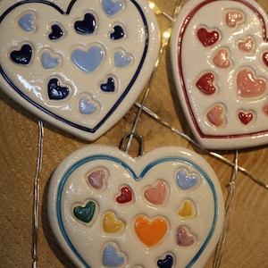 May include: Three heart-shaped ceramic ornaments with various coloured heart designs. The ornaments have a white base with blue, red, pink, yellow, and green hearts. Each ornament has a metal loop for hanging.