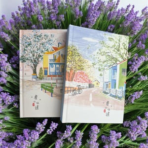 Może przedstawiać: Dwa notatniki w twardej oprawie z ilustracjami krajobrazów domów i drzew. Jeden notatnik przedstawia żółty i niebieski dom, zieloną ławkę i tekst "夏の小道". Drugi notatnik przedstawia zielony i niebieski dom, rower i ten sam tekst.