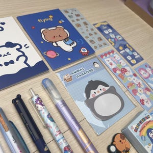 May include: A selection of stationery items, including pens, notebooks, and stickers. The items feature cute illustrations of animals, space themes, and other playful designs. The colour palette includes blues, purples, and pastels, creating a cheerful aesthetic.