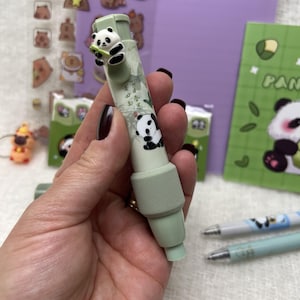 May include: A light green panda-themed pen featuring a panda figure and illustration. The pen has a light green grip and cap. Other panda-themed stationery items are visible in the background, including stickers and a notebook.