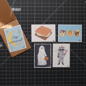 May include: A collection of whimsical Halloween-themed art prints. The prints feature a s'more, cookies with Halloween designs, a ghost holding a pumpkin, and a raccoon with a magic wand. One print has the text "Stay whimsical!" on a blue background.