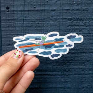 May include: A whimsical sticker featuring a cartoon frog rowing a boat through a sea of clouds. The frog is green and wearing a purple shirt. The boat is orange, and the clouds are shades of blue and white.