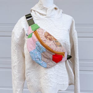 May include: A patchwork fanny pack featuring a variety of floral and solid-colored fabric hexagons in shades of pink, blue, tan, and red. The bag has a mustard yellow zipper closure and an adjustable olive green strap with a metal buckle.