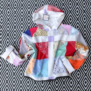 May include: A long-sleeve hooded sweatshirt constructed from a patchwork of various colorful fabric scraps including red, blue, orange, and floral patterns. The garment features a white hood with a tag reading Coco Clover and a black dotted hem.