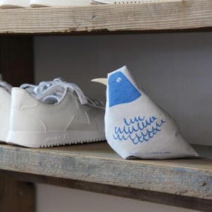 May include: A decorative fabric bird doorstop in white, with a blue head and wing pattern. The bird has a cream beak and sits on a wooden shelf next to white trainers.