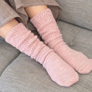 以下が含まれることがあります： ふくらはぎ上部に波状のテクスチャーのあるリブ編み模様が施され、足元は滑らかな平編み仕上げになっているライトピンクのニットソックス。丈はふくらはぎの中ほどまであり、全体的に柔らかく霜降りのような風合いが特徴です。