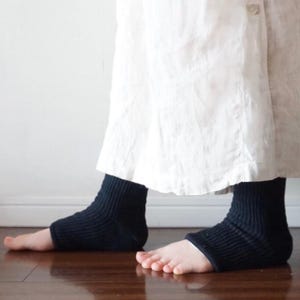 Könnte beinhalten: Ein Paar schwarze, gerippte Knöchelsocken, getragen zu einem weißen Rock. Die Socken haben ein dickes, strukturiertes Strickmuster, das den Fersen- und Mittelfußbereich bedeckt und die Zehen frei lässt. Der weiße Rock fällt locker über den Knöcheln.