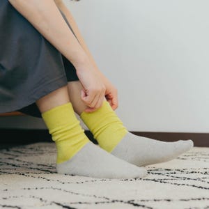 Puede incluir: Un par de calcetines grises claros con la parte superior amarilla. Una persona se está ajustando los calcetines. Los calcetines están sobre una alfombra blanca con patrones negros. La persona lleva un vestido gris.