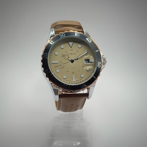 Op de afbeelding: Een automatisch Timex horloge met een beige wijzerplaat, zwarte bezel en bruine leren band. Het horloge heeft een zilveren kast en een datumvenster op 3 uur. De bezel heeft minutenmarkeringen.