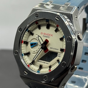 May include: A G-Shock Casio watch with a silver case and a light blue strap. The watch face is off-white with black, red, and blue accents. The watch displays the text "G-SHOCK CASIO" and "WR 20BAR".