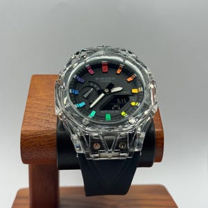 Puede incluir: Un reloj G-Shock Casio con caja transparente y esfera negra. El reloj tiene una correa negra y detalles de colores del arcoíris. El reloj se exhibe en un soporte de madera.