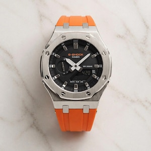 May include: A G-Shock Casio watch with a black dial and silver-tone case. The watch features an orange silicone band. The watch face displays the time as 4:20. The watch is water resistant to 20 bar.