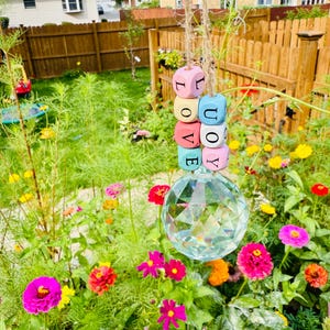 May include: A hanging garden ornament featuring wooden cubes spelling "LOVE" in pastel colors, suspended above a faceted crystal ball. The ornament hangs from a natural twine cord, set against a backdrop of a vibrant flower garden.