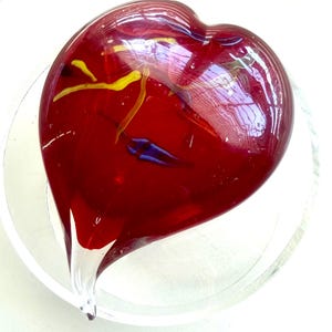 May include: A heart-shaped glass sculpture in shades of red, yellow, and blue, resting in a clear glass dish. The red glass heart has a glossy finish with streaks of yellow and blue. The dish is clear and circular.