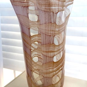 May include: A tall glass vase featuring a clear base and a complex, woven-like pattern of translucent pink and amber-colored glass ribbons. The design creates a series of open, oval-shaped gaps throughout the body, ending in a wide, flared rim.