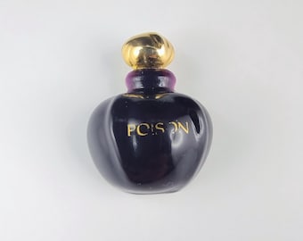Può includere: Un flacone di profumo viola scuro con un tappo color oro e la scritta "POISON" in lettere dorate. Il flacone ha una forma arrotondata e un collo viola. Lo sfondo è bianco.