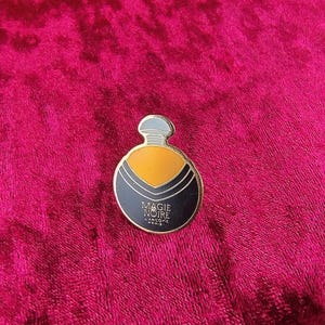 Puede incluir: Un broche de esmalte vintage con forma de frasco de perfume. El broche presenta un diseño negro y dorado con las palabras "MAGIE NOIRE" y "LANCOME PARIS" en letras blancas. El broche tiene una parte superior plateada y está sobre un fondo rojo.
