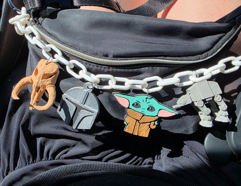 K&ouml;nnte beinhalten: Eine schwarze Bauchtasche mit einer wei&szlig;en Kette und mehreren Star-Wars-Anh&auml;ngern. Zu den Anh&auml;ngern geh&ouml;ren ein Sch&auml;del, ein Mandalorianer-Helm, Grogu und ein AT-AT-Walker. Die Anh&auml;nger sind in Braun-, Grau- und Gr&uuml;nt&ouml;nen gehalten.