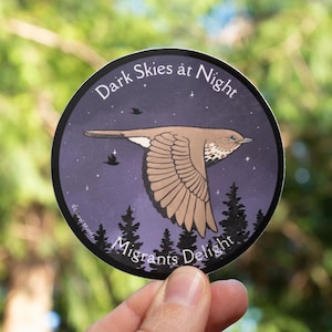 May include: A round sticker featuring a bird in flight against a dark purple night sky with stars and silhouettes of trees. The text "Dark Skies at Night" and "Migrants Delight" are printed on the sticker.