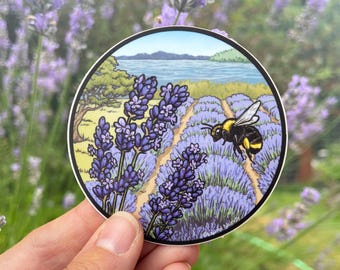 May include: A round sticker with a cartoon illustration of a bumblebee flying over a field of lavender. The background shows a lake and trees. The sticker has a black border and the text "Carol Mundy" at the bottom.