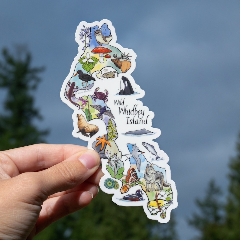 May include: A sticker featuring a map of Whidbey Island, Washington, with illustrations of local wildlife. The sticker includes a deer, orca, wolf, and other animals, with the text "Wild Whidbey Island". The sticker is held against a blurred background.