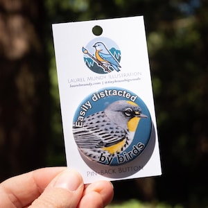 Puede incluir: Un botón redondo con una ilustración de un pájaro y el texto "Easily distracted by birds". El botón está sujeto a una tarjeta blanca con los datos del artista. El pájaro tiene marcas grises, amarillas y negras sobre un fondo azul.