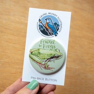 May include: A green tree frog pin-back button with the text "Friend to Frogs" on a light green background. The button is packaged with a white card featuring a bluebird illustration and the artist's name and website.