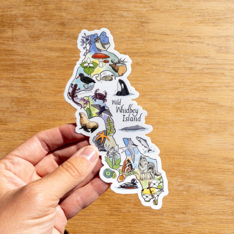 May include: A colorful sticker in the shape of Whidbey Island, featuring illustrations of local wildlife and flora. The sticker includes the text "Wild Whidbey Island" and various animal and plant illustrations.