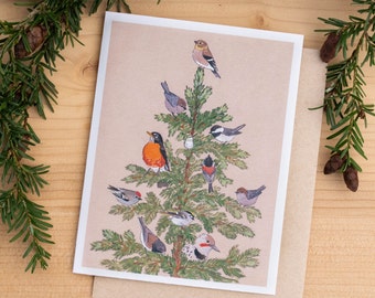 May include: A Christmas card with a watercolour illustration of a Christmas tree adorned with birds. The card sits on a wooden surface, with evergreen branches and pine cones. The card has a white border and a brown envelope.