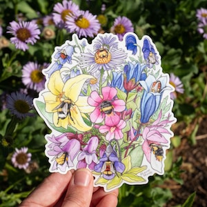 May include: A colorful sticker featuring a watercolor illustration of a bouquet of flowers with bees. The flowers are in shades of yellow, pink, purple, and blue. The sticker is held in front of a background of green foliage and purple flowers.