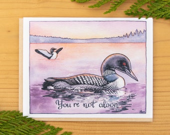 May include: A watercolor painting of two loon birds on a lake at sunset. The text "You're not alone" is written below the birds.