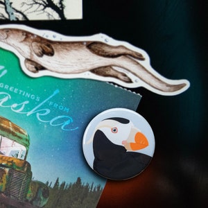 May include: A white and orange puffin with a black head and beak, illustrated on a round, white button.