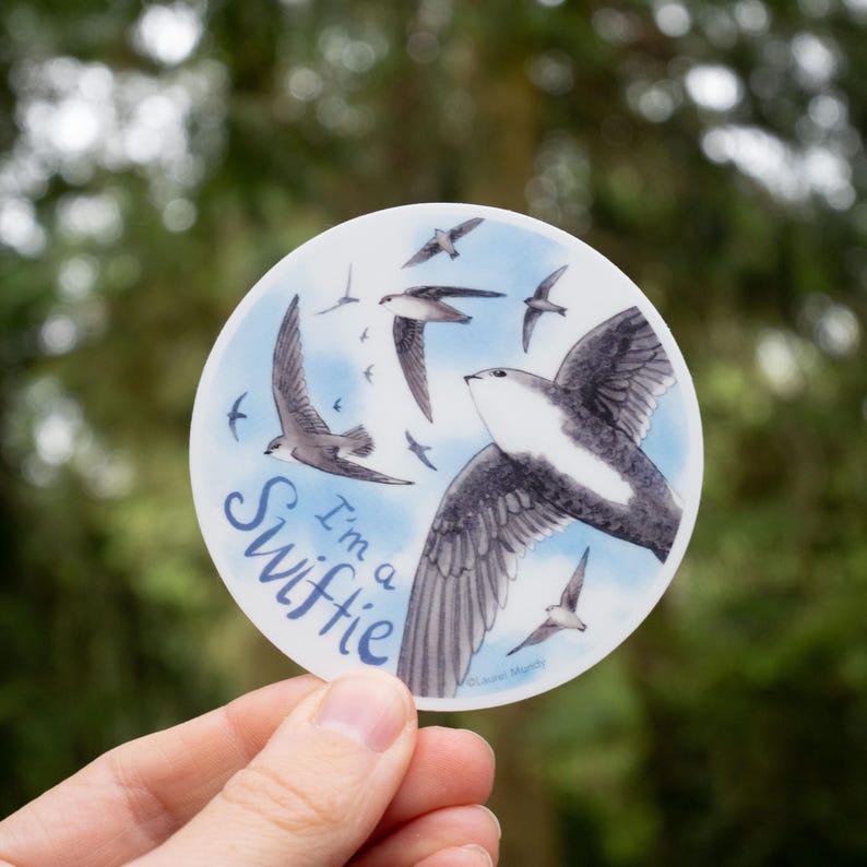 May include: A round sticker with a blue background and white clouds. The sticker features a black and white illustration of a swift bird in flight with the text "I'm a Swiftie" in a stylized font.