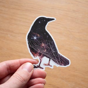 May include: A black crow silhouette sticker with a galaxy pattern inside. The sticker features a starry night sky with a bright star in the center.