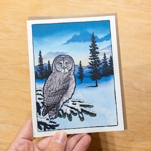 May include: A watercolor illustration of a gray owl perched on a snowy branch in a wintery forest scene. The owl has yellow eyes and is looking directly at the viewer. The background is a blue sky with clouds and snow-covered trees.