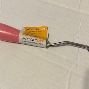 May include: A pink-handled latch hook tool with a silver metal hook. The tool has a yellow and white label that reads "wonderart" and "Latch Hook Tool". The tool is designed for crochet and other crafts.