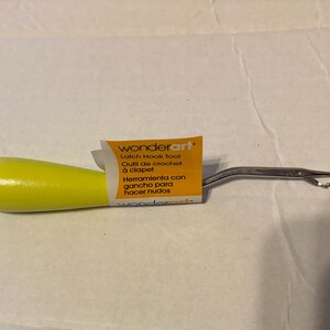 May include: A yellow-handled latch hook tool with a metal hook end. The tool has a label that reads "wonderart" and includes text in English, French, and Spanish. This tool is used for latch hook crafts.