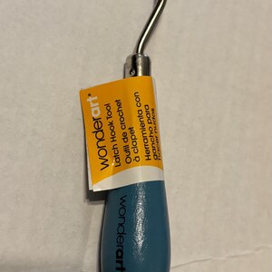 May include: A Wonderart latch hook tool with a blue handle and a silver hook. The tool has a label with text in multiple languages, including "Latch Hook Tool" and "Outil de crochet à clapet."