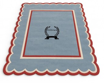 May include: A rectangular rug with a scalloped edge. The rug features a light blue center, a red border, and a cream-colored inner border. A black emblem with text is in the center of the rug.