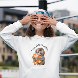 Può includere: Felpa con cappuccio bianca con una grafica di due scimmie che si abbracciano. Il testo "found comfort in love..." è sopra l'immagine. La felpa ha una tasca frontale e un cappuccio con coulisse.