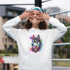 May include: White hoodie featuring a colorful wolf design. The wolf's portrait is rendered in vibrant hues of blue, purple, yellow, and pink, with a splash art effect. The hoodie has a front pocket and a drawstring hood.