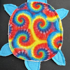 May include: A turtle-shaped fabric item featuring a vibrant tie-dye shell pattern in red, yellow, and blue spirals. The head and four flippers are solid light blue fabric with visible stitching. Two small black googly eyes are attached to the head.